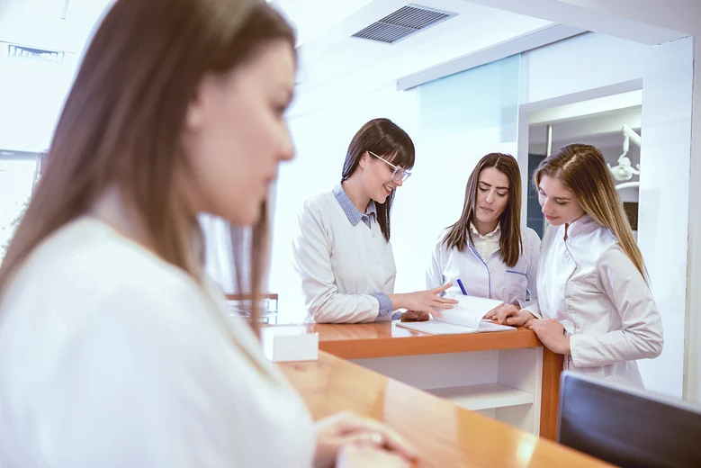 Medizinische Fachangestellte in Arztpraxis besprechen Unterlagen am Empfang. MFA organisieren Praxisabläufe, empfangen Patienten, koordinieren Termine, führen Abrechnungen durch und assistieren bei Untersuchungen sowie Behandlungen im ärztlichen Team.